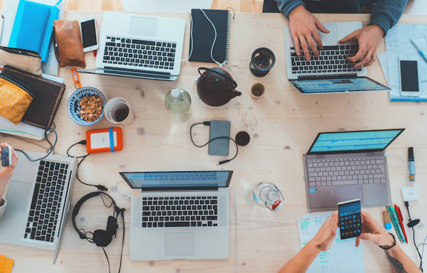 Overhead view of a wooden table with laptops, phones, cables, a teapot and cups
