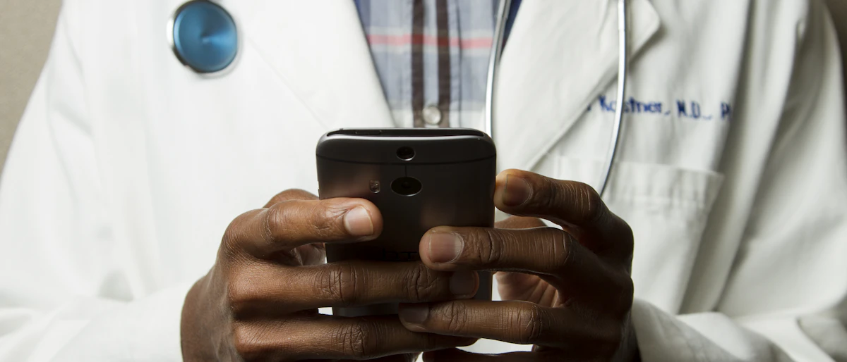 Beyaz önlüklü ve boynunda stetoskop olan bir kişi iki eliyle akıllı telefon kullanıyor